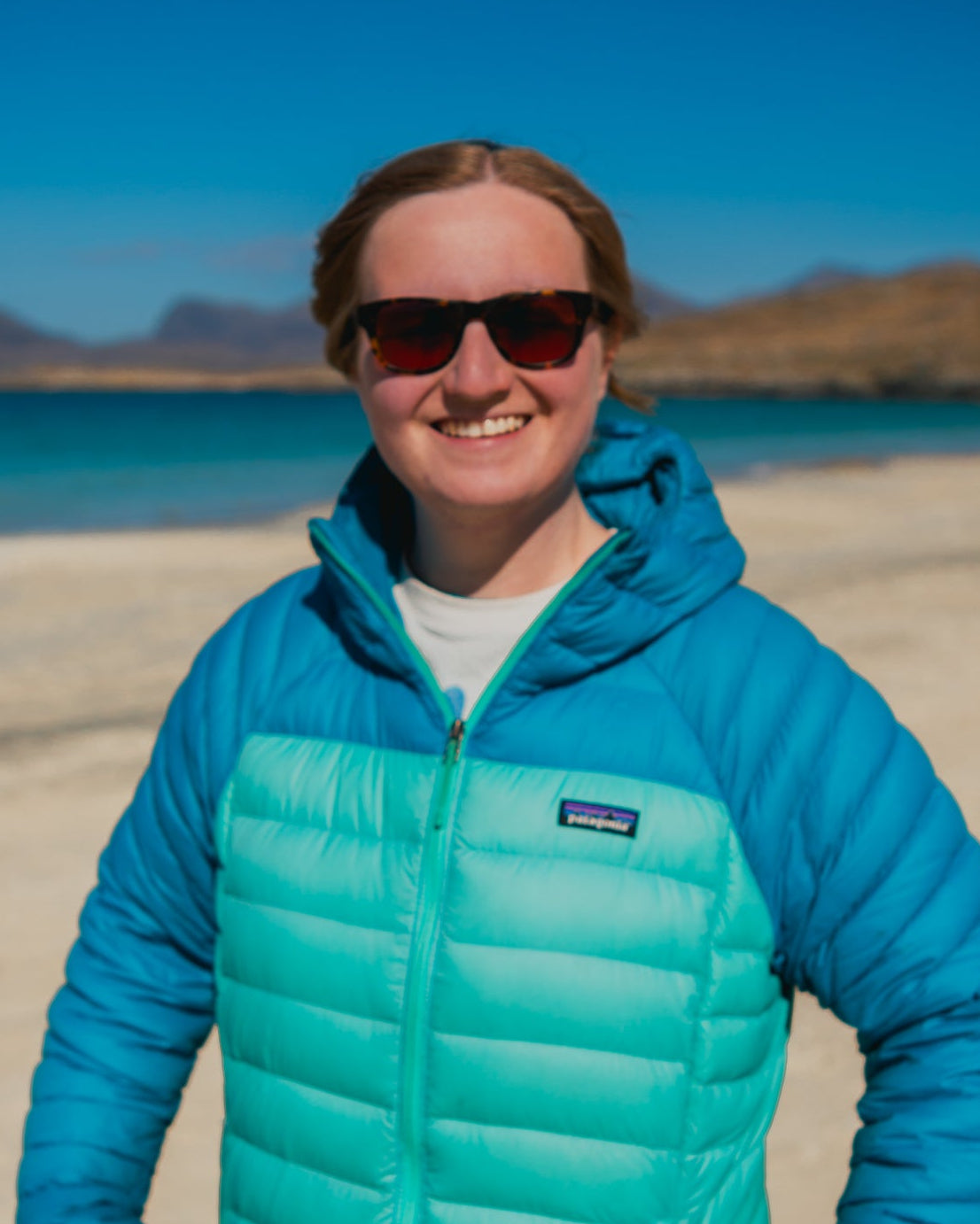 Person wearing a blue and green jacket with a Patagonia logo on a beach with mountains and water in the background.