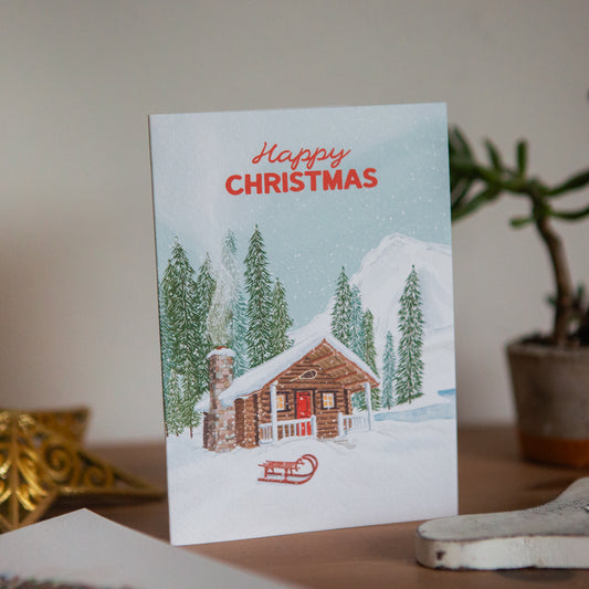 Christmas card with a snowy cabin and trees on a table.
