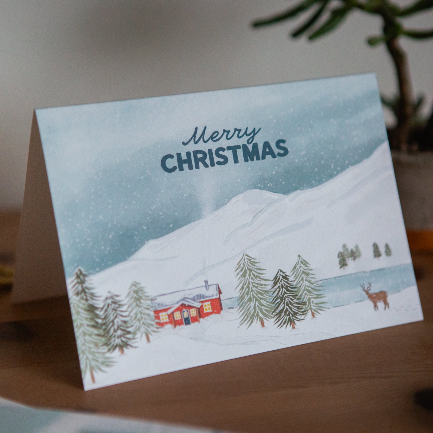 Christmas card with a snowy landscape, cabin, and deer on a wooden surface.